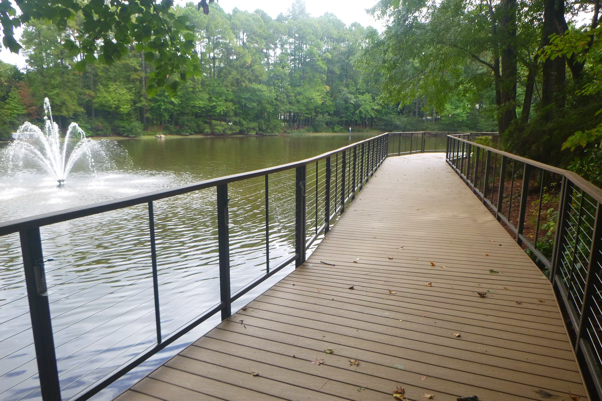 A composite bridge with metal rails over water with a fountain in the background.